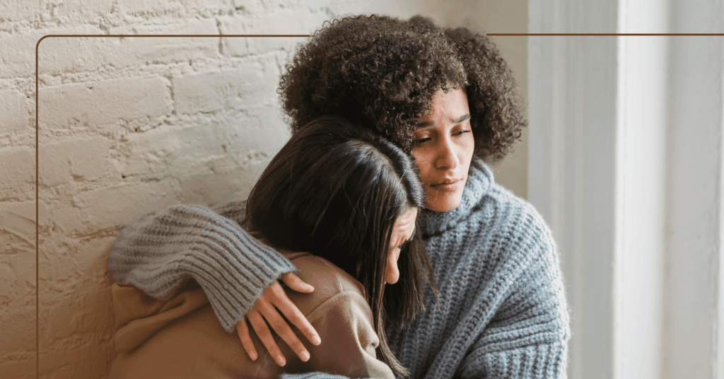 Two women hugging. One woman is wearing a fluffy gray sweater and the other is in a brown shirt. They are leaning against a white brick wall.