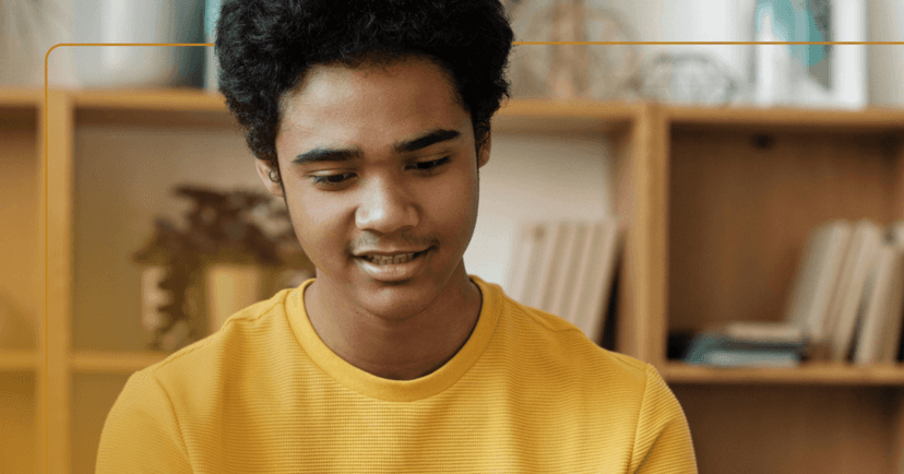 Young man sits staring down at the floor in front of him. His teeth are visible as if he is about to speak. He is wearing a yellow long sleeve shirt. The background is a tan bookshelf.