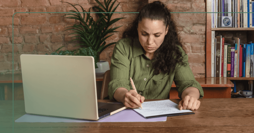 Woman sits at a desk with an open laptop in front of her. She is looking down at the notes she is writing with a pen in her right hand. She is wearing a green shirt and her facial expression is focused. The background is a green plant against a brick wall and a bookshelf.