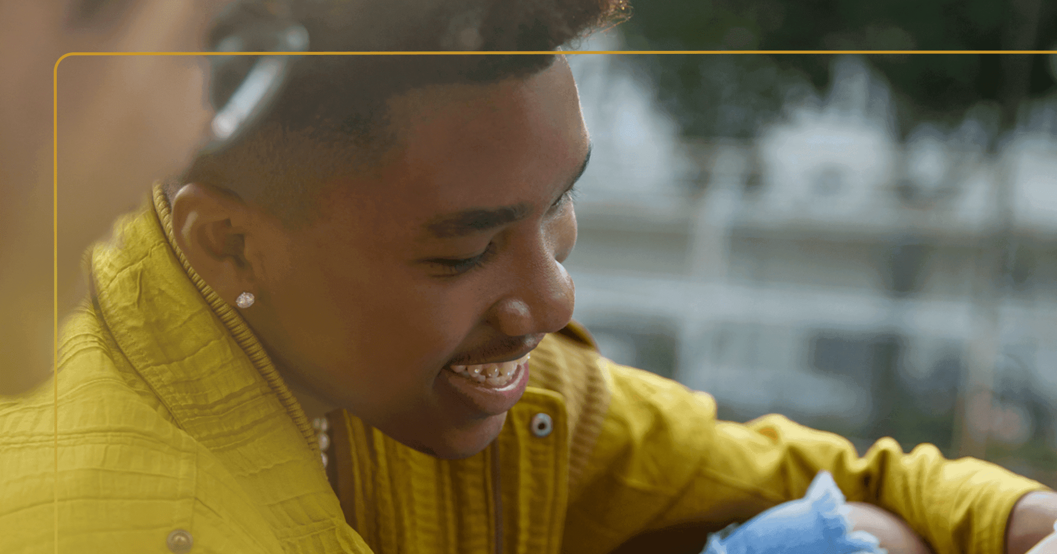 Young man sits with arms around his knees, looking downward. He is smiling. He's wearing a yellow jacket and jeans ripped at the knee. The background is blurry, and he appears to be outside.