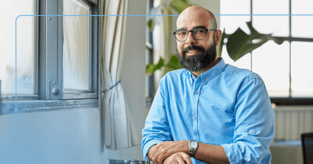 Man stands leaning on a table near a window. He is staring straight at the camera and smiling with his teeth. He is wearing a blue button-up shirt and black round glasses. The background is a blurry plant and six windows.