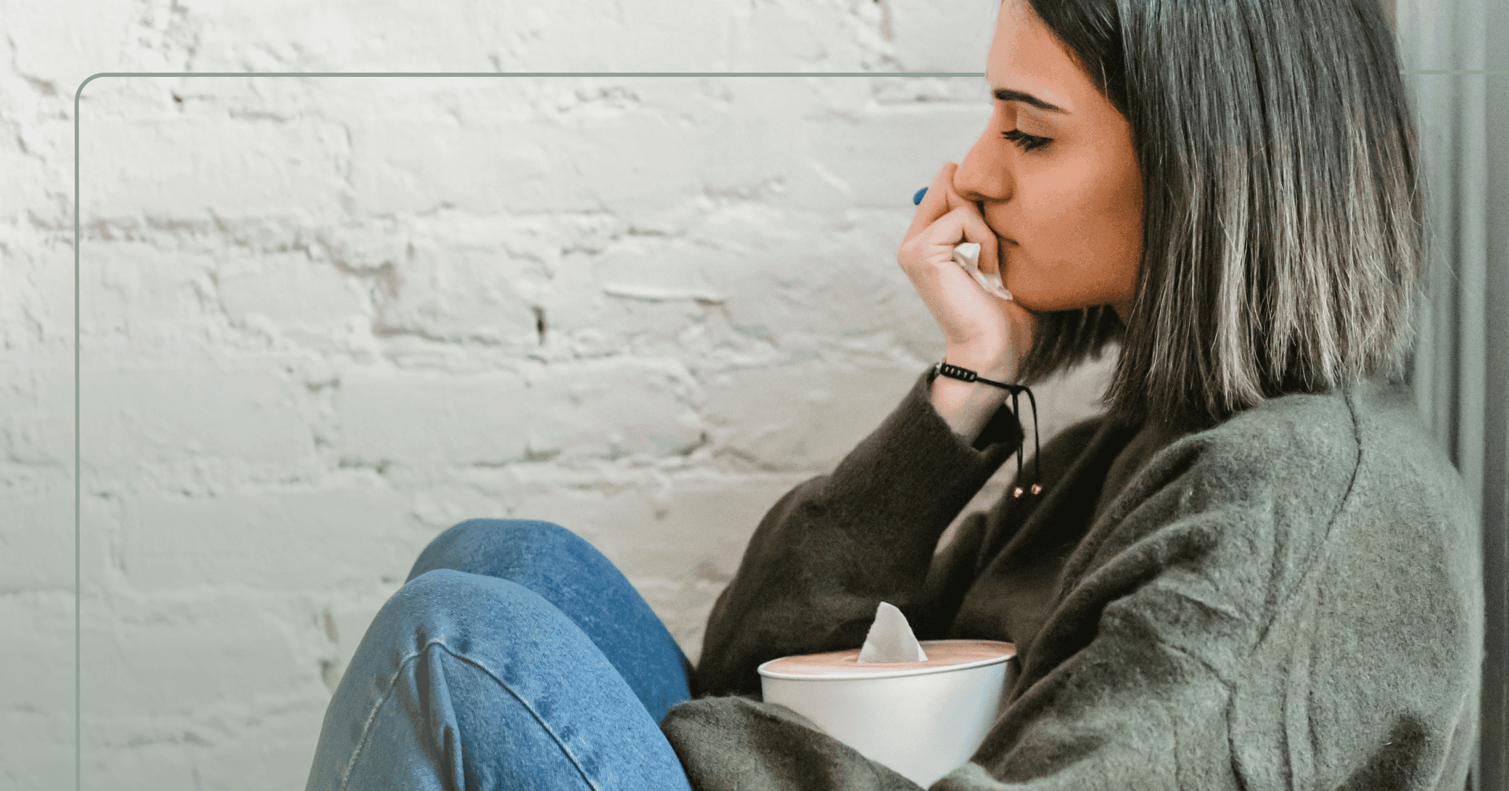 A woman is holding a tissue box with a sad look on her face. She is wearing a green sweater and sitting in front of a white brick wall.