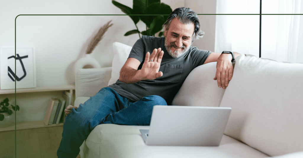 Older man sits on a white couch with an open laptop in front of him. He is waving his right hand and smiling looking down at the laptop screen. He is wearing a dark green T-shirt and jeans. The background is a bookshelf against a white wall and a window to the right.