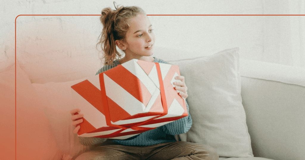 Young girls sits on a couch and holds a large present wrapped in red and white striped wrapping paper. The image is framed with a red line.