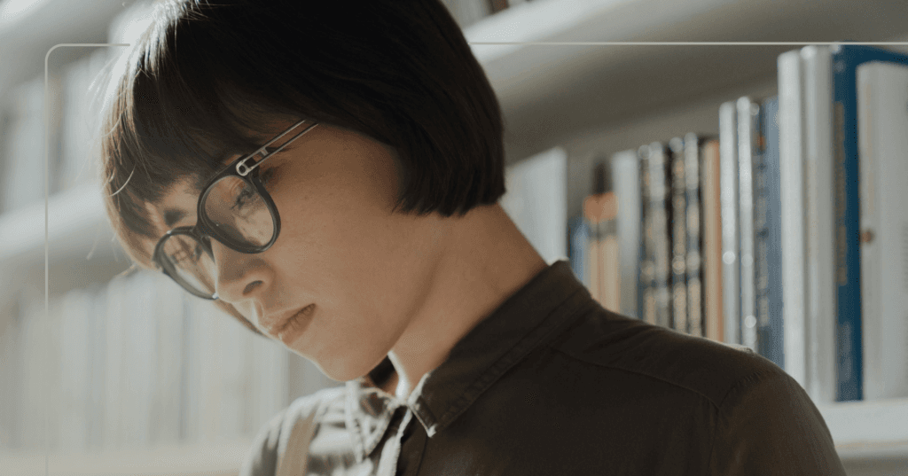 Woman stands in front of a book shelf. She is looking down at something in front of her offscreen. She is wearing black round glasses and a brown button-up shirt. The background is hardcover books that are blurry.