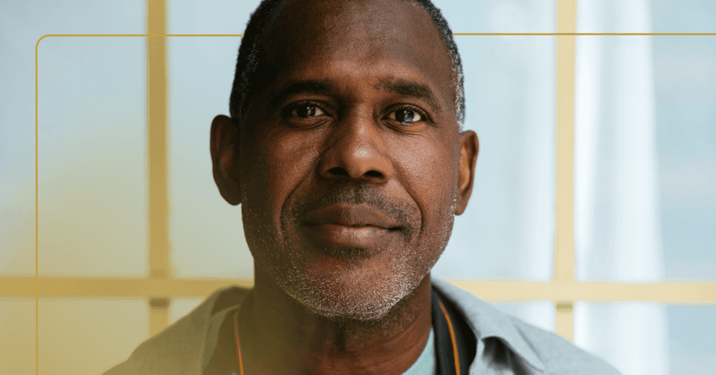 Man stands staring straight at the camera. His facial expression is calm and invested. He is wearing a gray button up shirt. The background is a light blue sliding curtain.