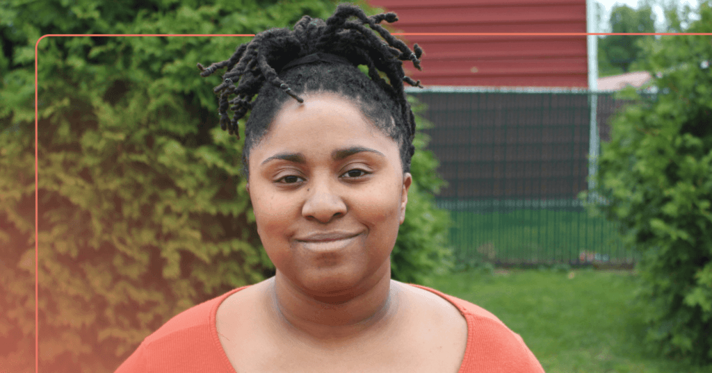 Woman stands outside staring straight at the camera. She is smiling. She is wearing an orange long sleeve shirt. The background is a backyard with trees.