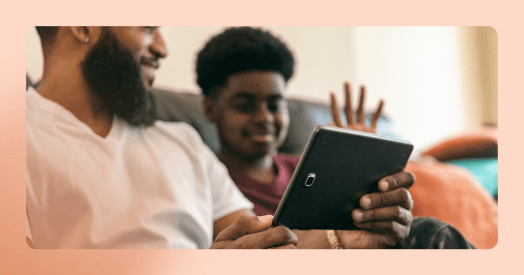 Father and son sit on the couch together with Dad holding a tablet in his hands. The son is waving at someone possibly on a video call on the tablet. Dad is wearing a white V-neck shirt and son is wearing a red T-shirt. The background is a white wall.