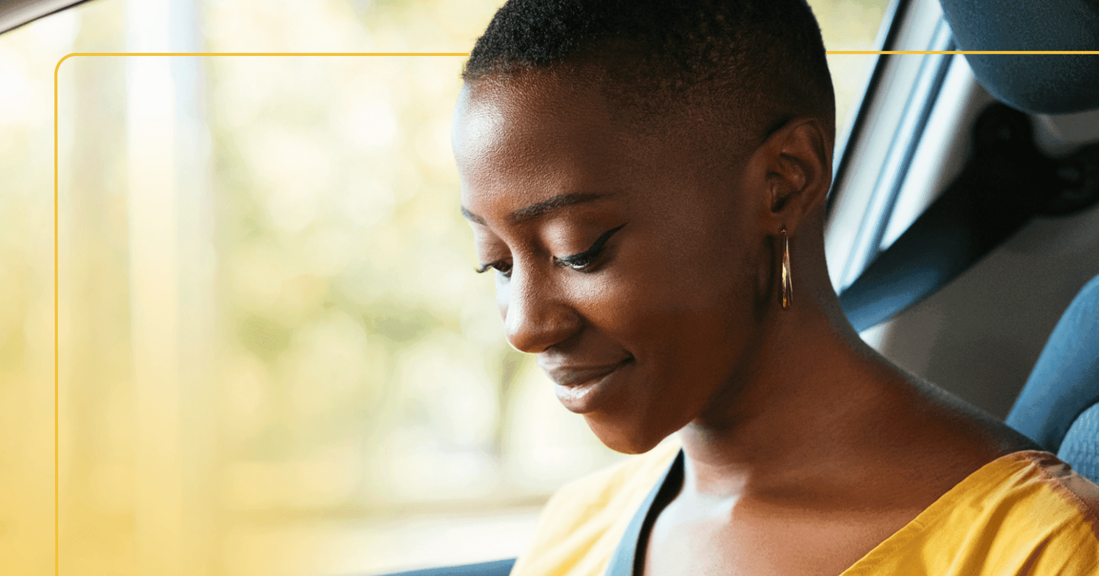 Woman sits in car with seatbelt on. She's looking down, possibly at a phone. She's wearing a yellow shirt and the background outside of the window is green and yellow.