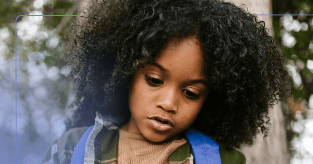 Child is in the center of the image staring down in front of them. Their mouth is slightly open gazing at something offscreen. They are wearing a tan shirt underneath a green flannel with blue backpack straps visible. The background is trees.
