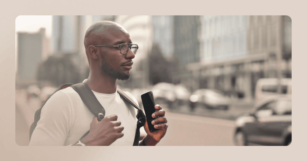 A man wearing a backpack and holding a smartphone looks off camera to the right. He is standing on a city block and wearing glasses.