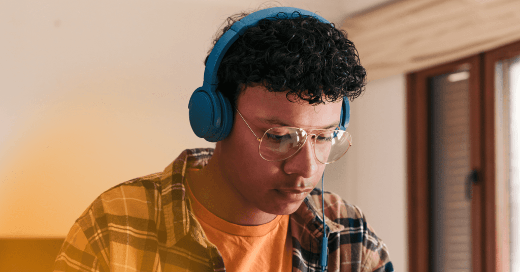 Man sits wearing blue over the ear headphones looking down at something offscreen. He is wearing an orange T-shirt underneath a flannel shirt and glasses. The background is a white wall.