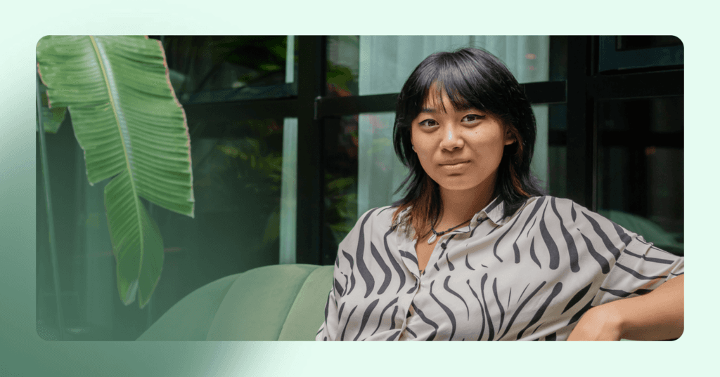 Woman sits on a couch staring straight into the camera. She has her left arm propped up and has a slight smile. She is wearing a white and black patterned shirt and a necklace. The background is glass windows.